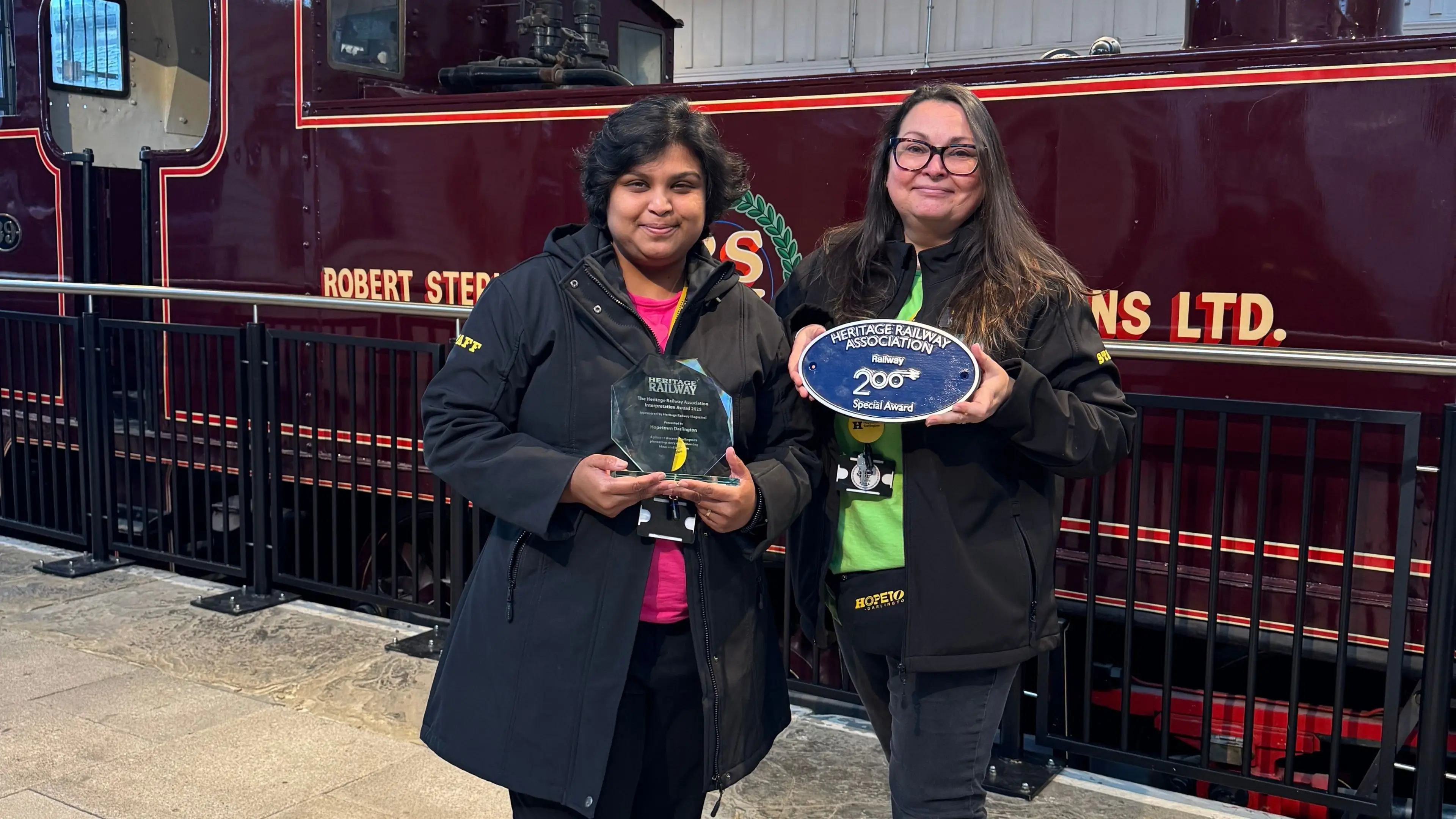Two Hopetown female staff members stood in front of Locomotive No.39 holding two awards